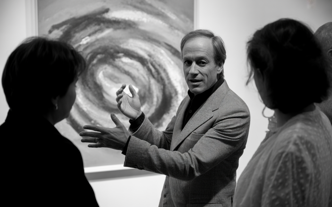 Un homme en costume gris explique une œuvre d'art abstraite à deux femmes, dans une galerie d'art.