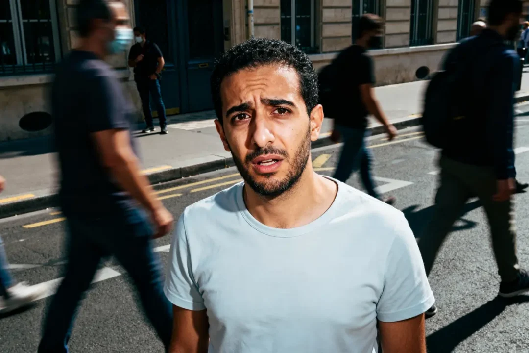 Un homme en t-shirt blanc sur un trottoir, entouré de passants flous.