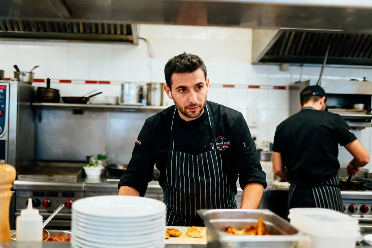 Chef en cuisine professionnelle, portant un tablier, devant un plan de travail avec assiettes et plats préparés.