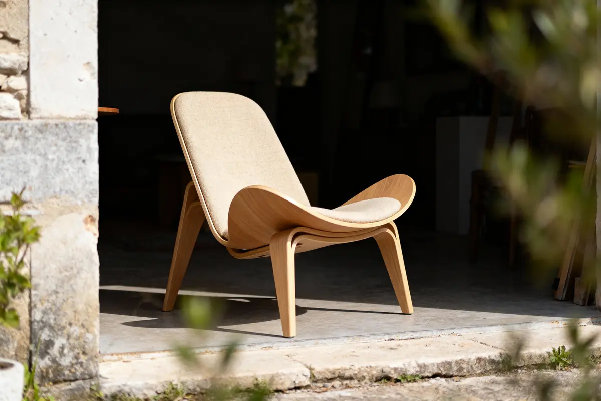 Chaise design en bois et tissu beige, éclairée par le soleil, située dans une entrée en pierre.