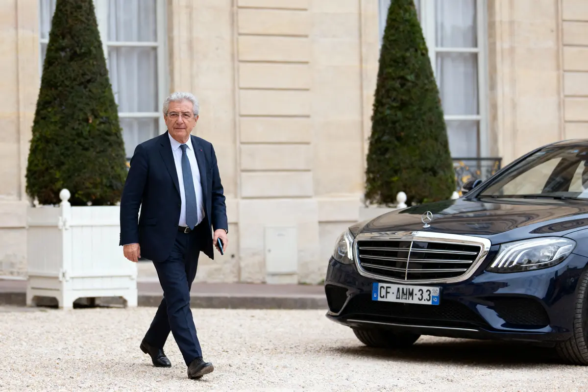 Homme en costume marche près d'une voiture Mercedes devant un bâtiment officiel avec des arbustes en arrière-plan.