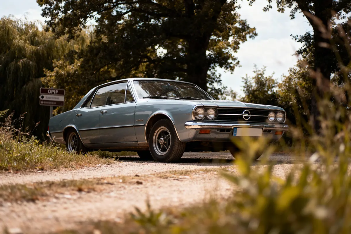 Voiture classique Opel grise garée sur chemin de terre, entourée d'arbres, ciel bleu en arrière-plan.