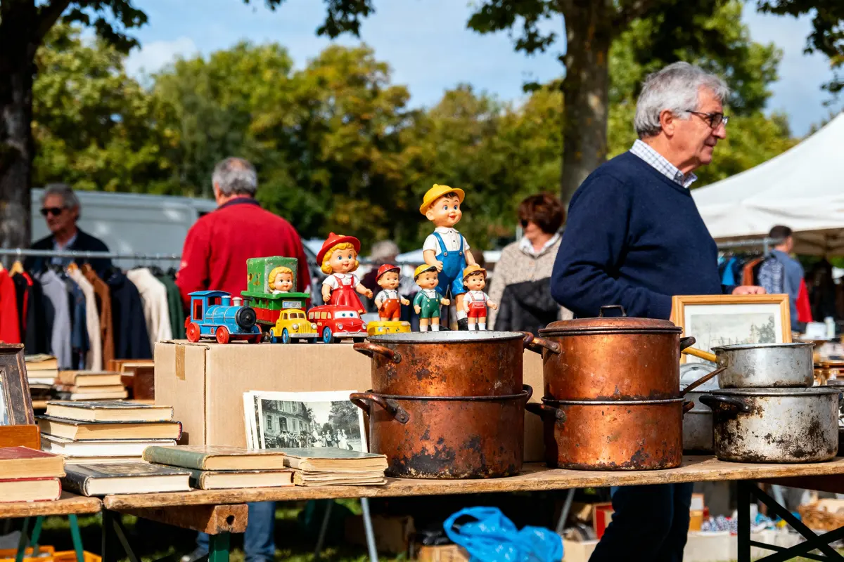 Stand de brocante avec jouets vintage, livres, casseroles en cuivre, et personnes observant les objets exposés.