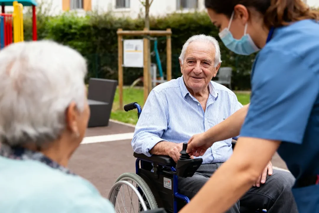 Personne âgée souriante en fauteuil roulant, assistée par une infirmière masquée, parlant à quelqu'un dehors.