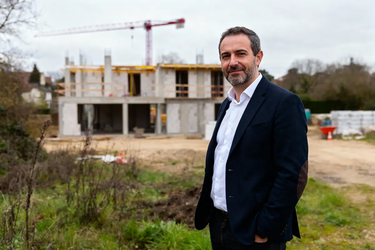 Homme en costume debout devant un chantier de construction avec grue en arrière-plan.
