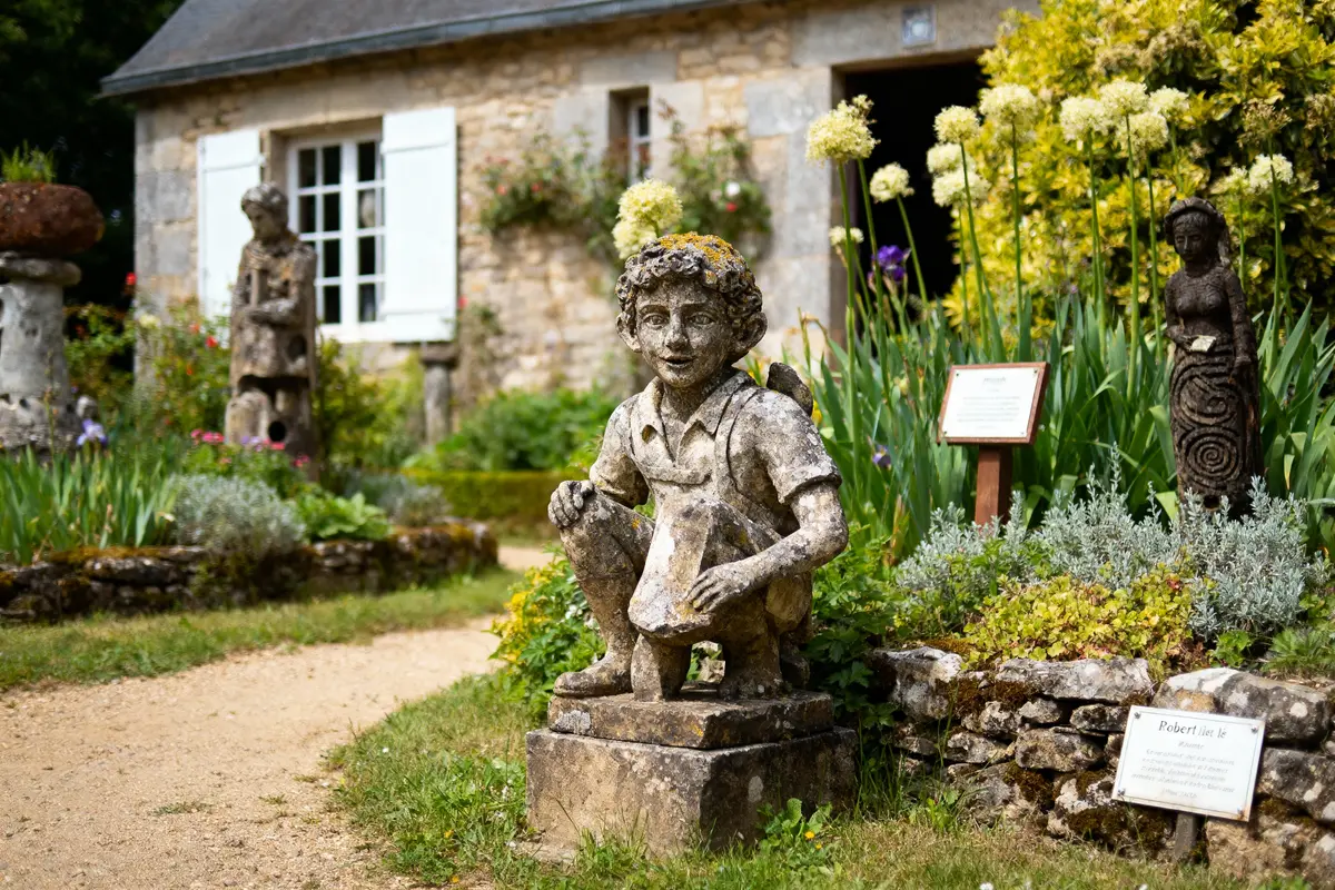 Statues en pierre dans un jardin fleuri devant une maison en pierre avec fenêtres blanches.