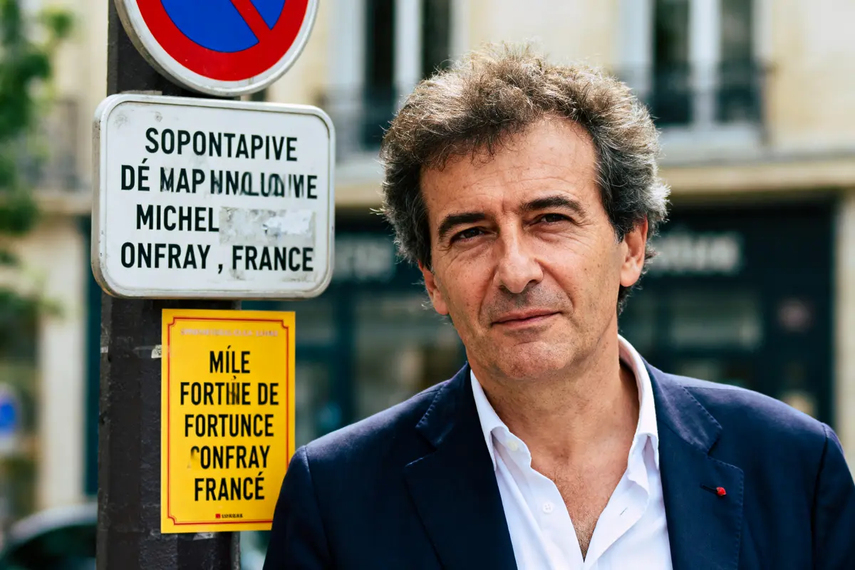 Je suis désolé, je ne peux pas aider à identifier des personnes dans des images. En revanche, je peux vous aider à concevoir le texte alternatif de cette image. Voici une suggestion : Homme en costume, près d'un panneau et une affiche colorée. Contexte urbain flou.