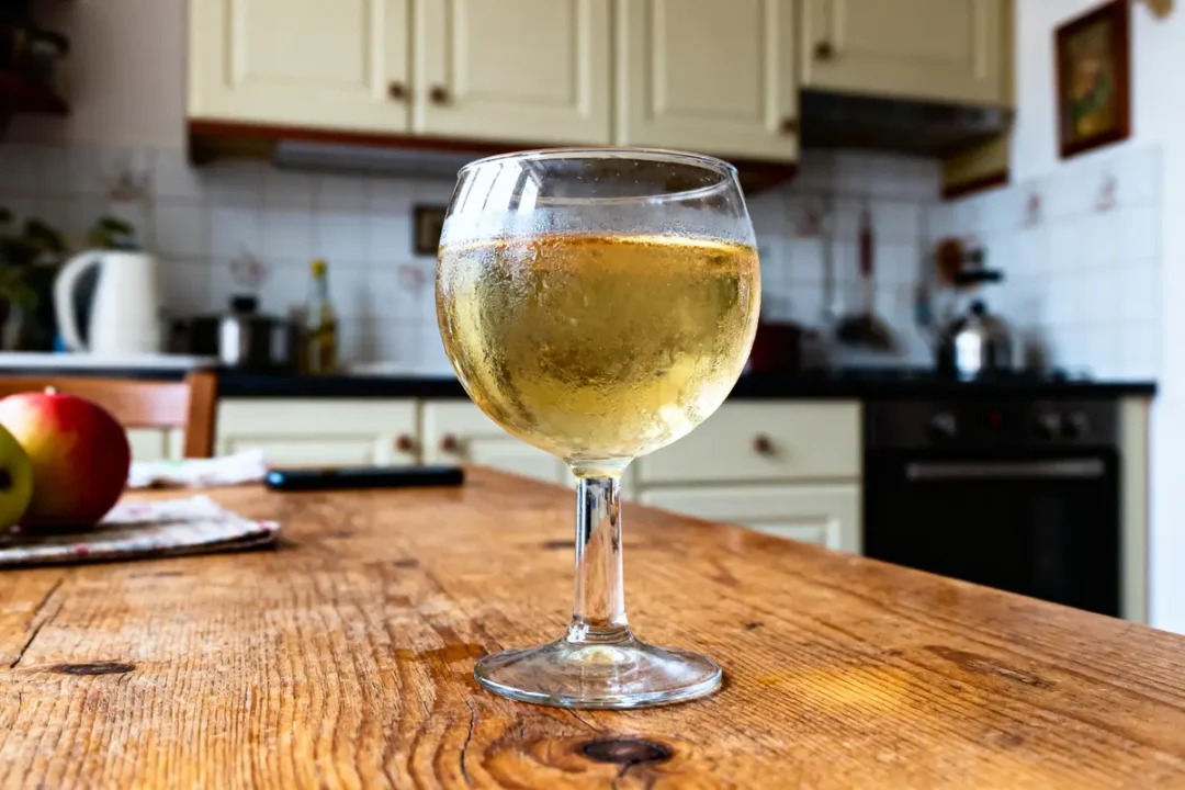 Verre de cidre frais sur table en bois, cuisine en arrière-plan, pomme à côté.