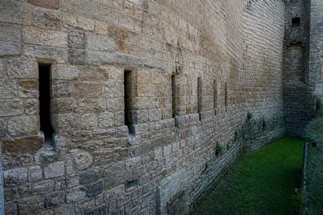 Fortifications médiévales avec meurtrières adjacentes et barbacane en pierre entourée de verdure.
